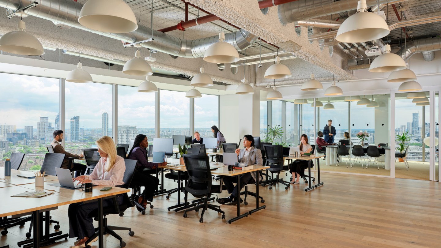 WeWork members doing focused work at their desks, while others hold a meeting in a separate room in the background