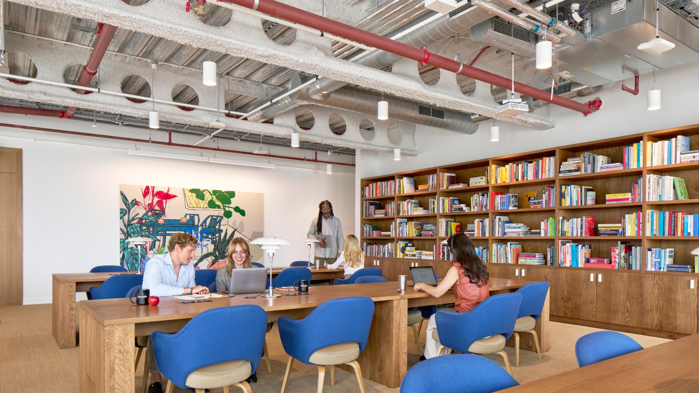 WeWork members sitting at shared desks in a colorful space, with books on shelves in the background