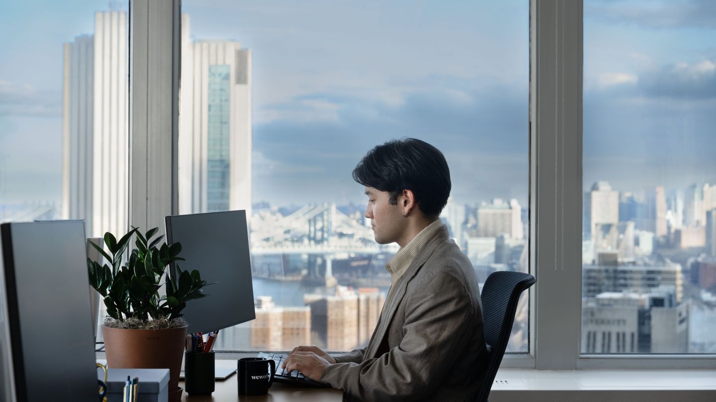 A person working sitting at their desk; large windows overlooking the cityscape in the background. 