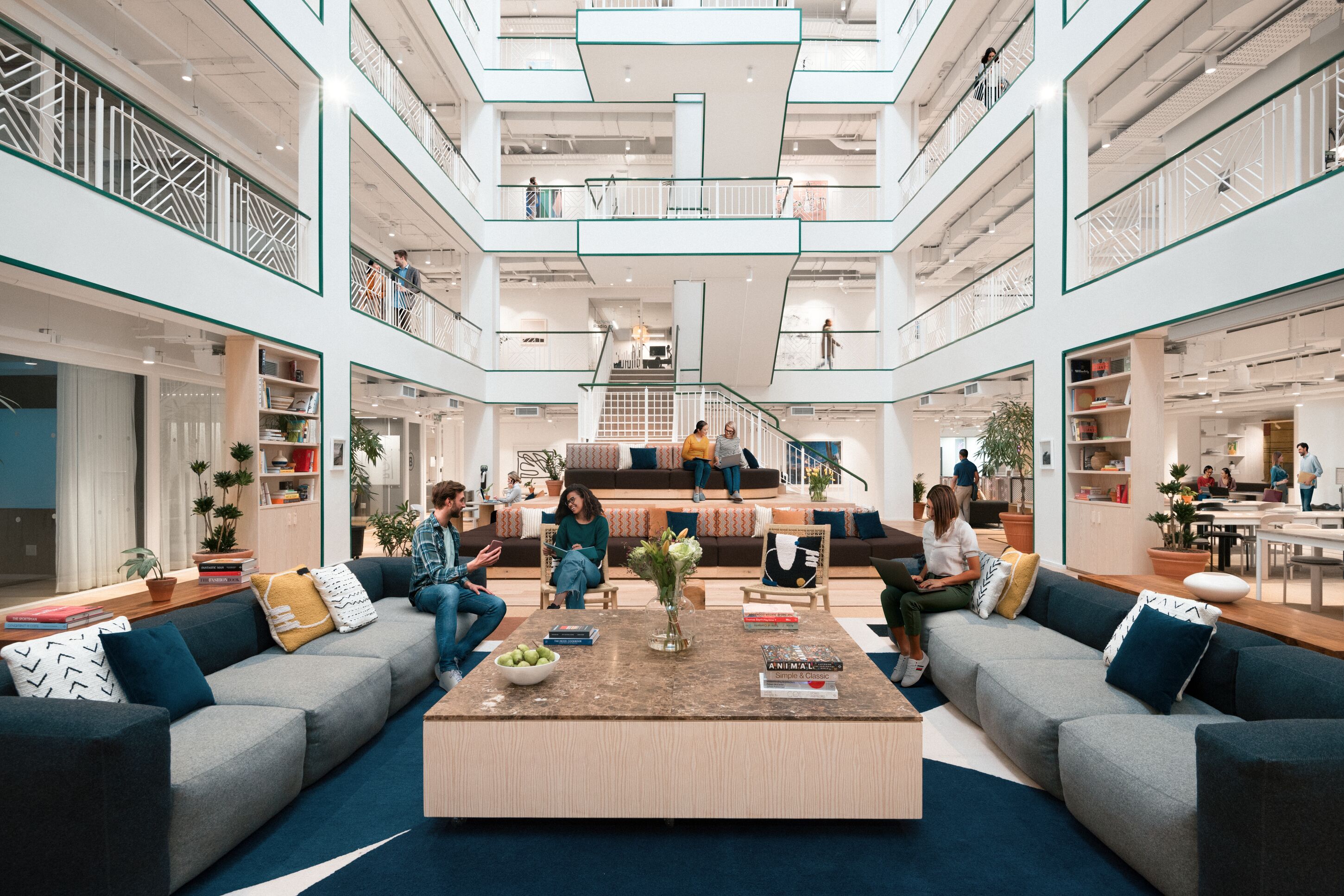 Modern multi-story office atrium where professionals sit on couches and tiered seating working on laptops and collaborating.