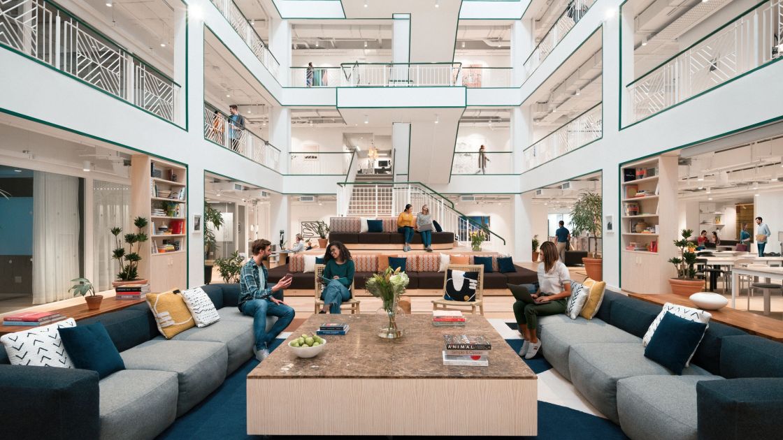 Modern multi-story office atrium where professionals sit on couches and tiered seating working on laptops and collaborating.
