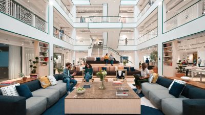 Modern multi-story office atrium where professionals sit on couches and tiered seating working on laptops and collaborating.